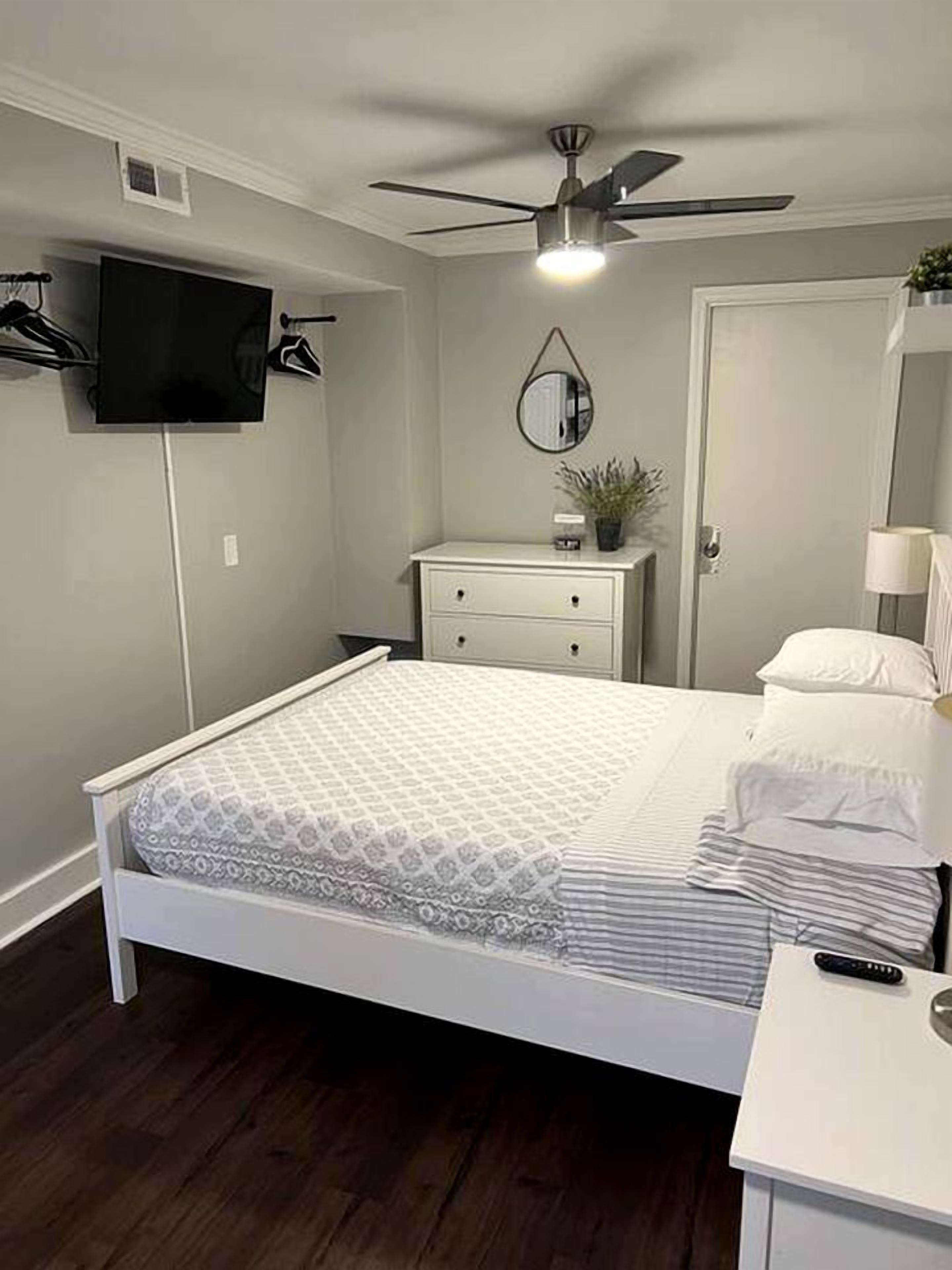A bedroom with a bed, a bedside table, a dresser, a ceiling fan, a TV mounted on the wall, and a door.