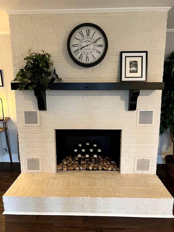 A white brick fireplace with a clock, a framed picture, and a plant.