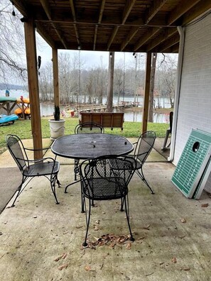 An outdoor patio with a round table and four chairs, a covered area with a wooden ceiling, and a view of a lake and dock area.