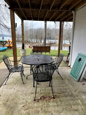 An outdoor patio with a round table and four chairs, a covered area with a wooden ceiling, and a view of a lake and dock area.