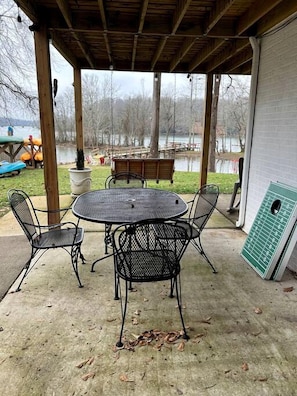 Une terrasse extérieure avec une table ronde et quatre chaises, un espace couvert avec un plafond en bois, et une vue sur un lac et une zone abritant un quai.