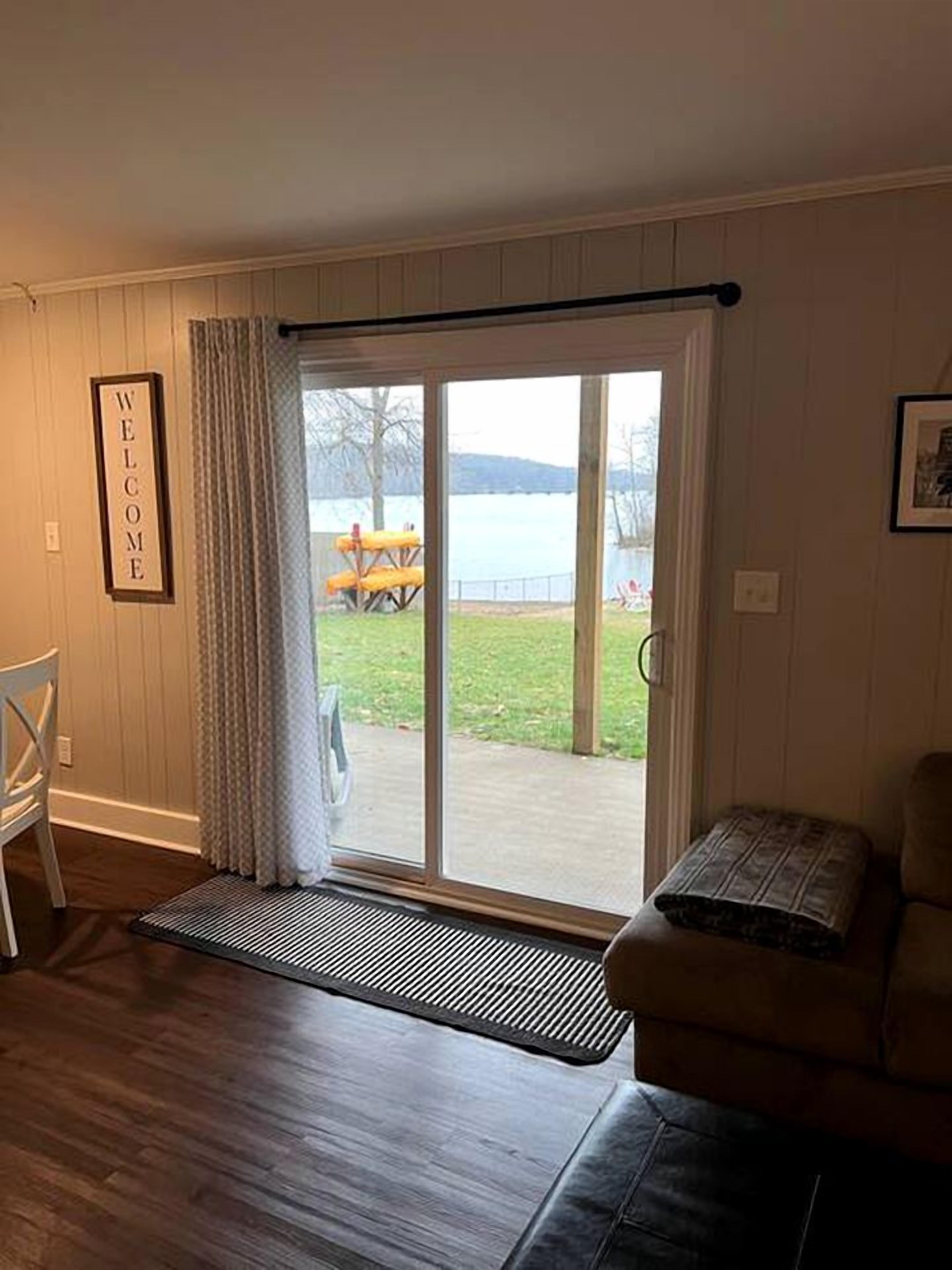 Una habitación con puerta de vidrio corredera que da a un patio con mesa y sillas, un cartel de bienvenida enmarcado y una zona de estar con cojines.