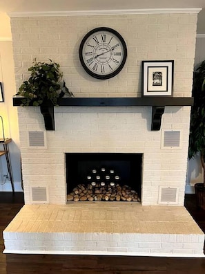 Une cheminée en briques blanches ornée d’une horloge, d’un tableau encadré et d’une plante.
