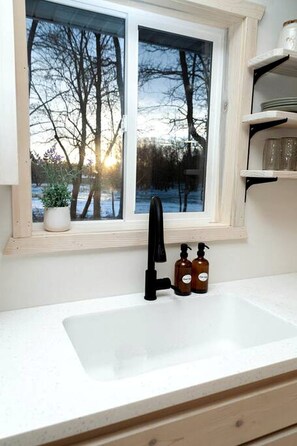 Una cocina con ventana, un lavamanos y una estantería con botellas.