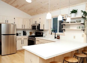 A modern kitchen with stainless steel appliances, wooden cabinets, a large island, pendant lights, and a window with a view.
