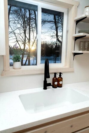 A kitchen with a window, a sink, and a shelf with bottles. - Cabin Charm by the Pine: Pet-Friendly Retreat in Mission Township's Riverside Haven (Mission Township)