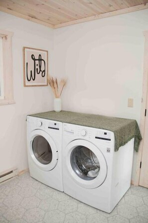 A laundry room with two white front-loading washing machines, a framed artwork on the wall, and a small vase with reeds on top of the machines.