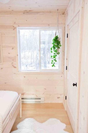 Una habitación de cabaña de madera con una cama, una ventana que ofrece vistas a árboles cubiertos de nieve y una planta colgante.