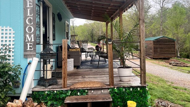 A blue building with a wooden porch, a welcome sign, outdoor furniture, and a shed in the background. - Sweet Tiny House Perfect for a Romantic Retreat in Alabama (Rogersville)