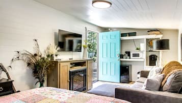 A bedroom with a bed, a dresser, a television, a blue door, and a kitchenette.