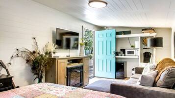 A bedroom with a bed, a dresser, a television, a blue door, and a kitchenette.