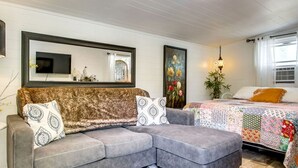 Een kamer met een grijze bank, een televisie, een schilderij, een bed met een kleurrijk sprei en een lamp.