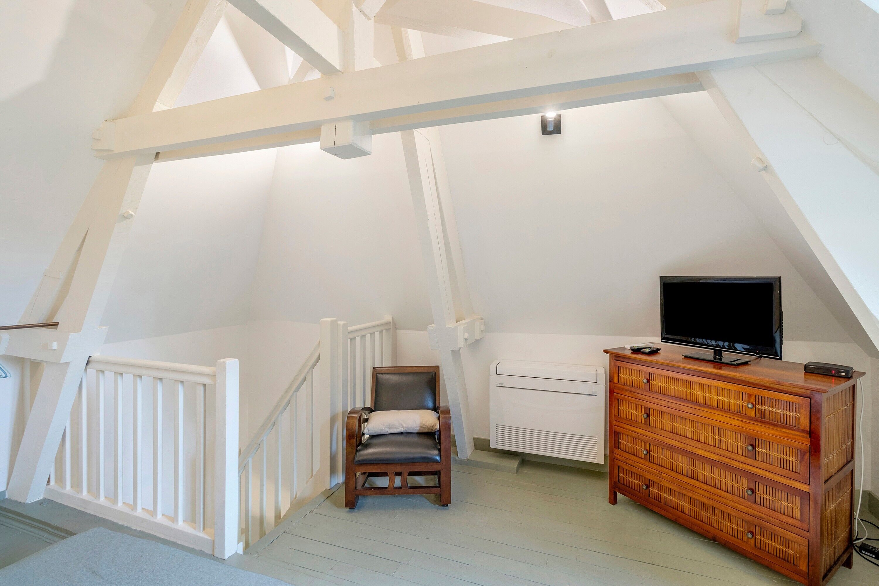 Une pièce avec un plafond en pente, une chaise en bois, un téléviseur posé sur une commode et un radiateur blanc.
