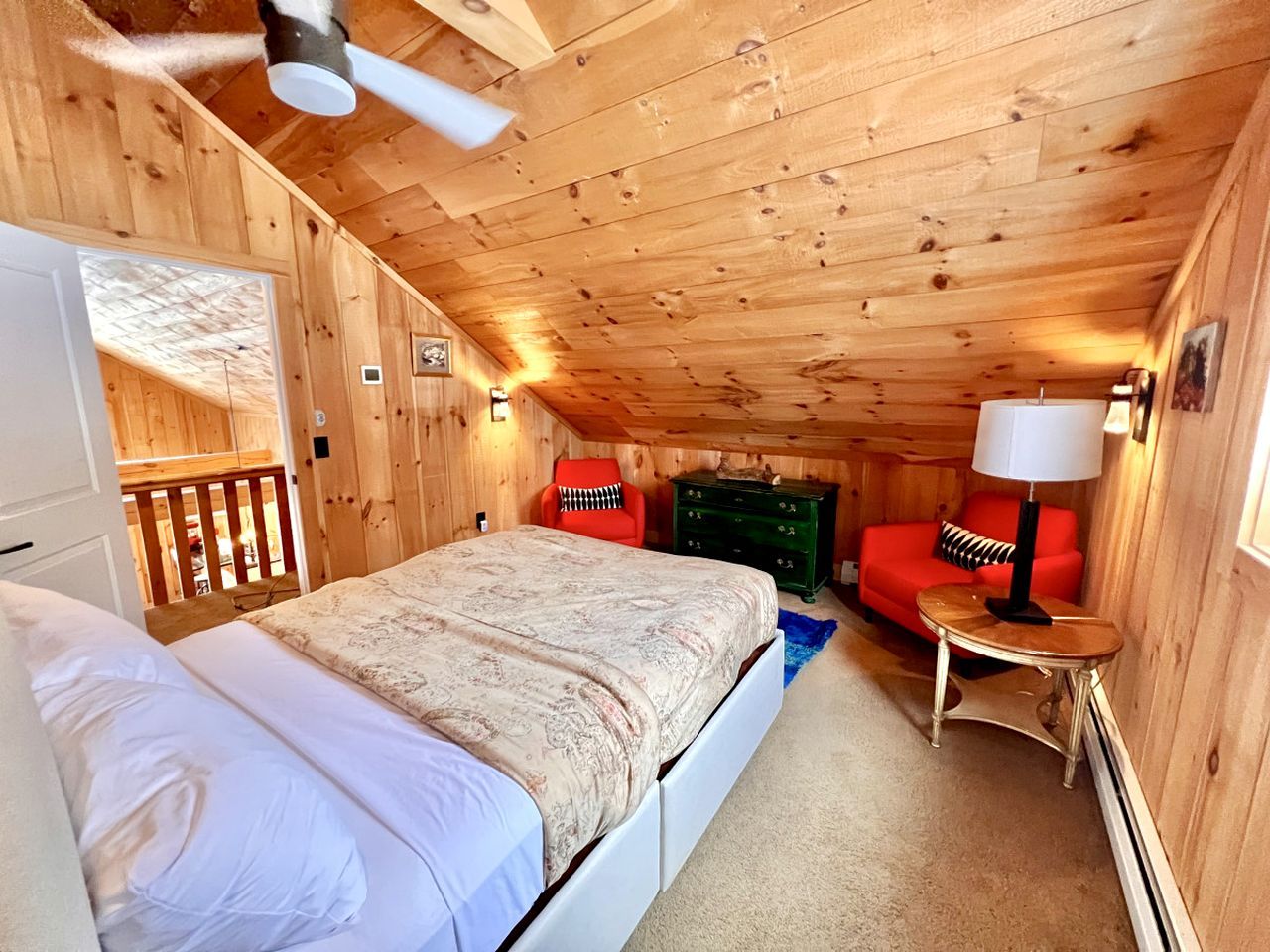 Une chambre dans une cabane en bois, avec un lit, une chaise rouge, une commode verte et un ventilateur de plafond.