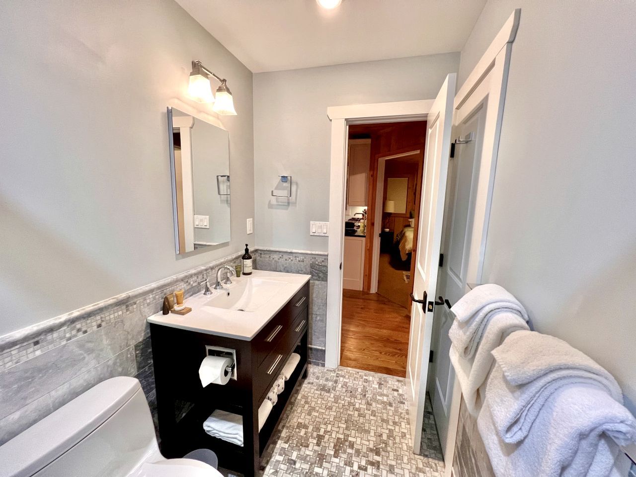 Une salle de bain avec des toilettes blanches, un lavabo avec un miroir, un porte-serviettes et un sol carrelé.