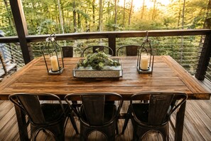 A wooden deck with a table set for four, surrounded by metal chairs, with a railing and forest view in the background.