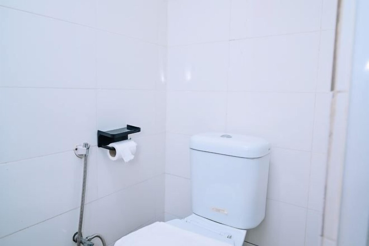 A small, white, tiled bathroom with a toilet, a paper towel holder, and a handheld showerhead.