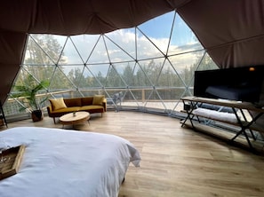 A spacious room with a large bed, a sofa, a round table, a TV on a wooden stand, and a view of trees through a geometric window structure. - Spacious Dome with Lofted Interior and Furnished Deck in Kentucky (Cadiz)