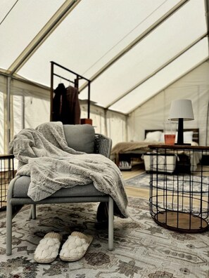 A tented room with a grey chair, a small table with a lamp, and a pair of slippers on the floor. - Remote Expansive Tented Cabin Incredible for a Glamping Experience in Nevada (Boulder City)