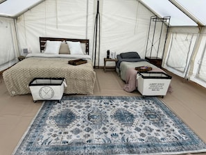 A tented accommodation with a bed, a small table, a chair, a rug, and a laundry service cart.