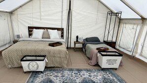 A tented accommodation with a bed, a small table, a chair, a rug, and a laundry service cart.