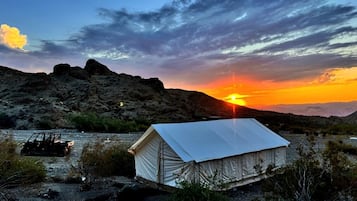A tented structure with a flat roof, surrounded by sparse vegetation and rocky terrain, with a sunset in the background.