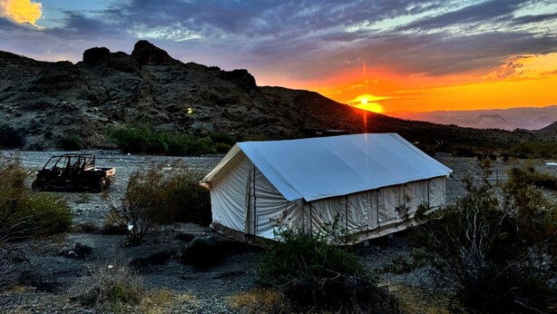 A tented structure with a flat roof, surrounded by sparse vegetation and rocky terrain, with a sunset in the background.