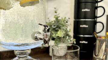 A glass water dispenser with a lemon slice, a stack of black coffee mugs, and a small glass on a wooden surface.