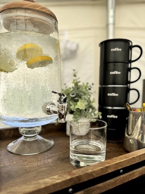 A glass water dispenser with a lemon slice, a stack of black coffee mugs, and a small glass on a wooden surface.