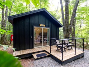Una pequeña cabaña negra con puerta de vidrio, una terraza con asientos exteriores y un cartel que dice 'CAN'T BE LEAVING'.