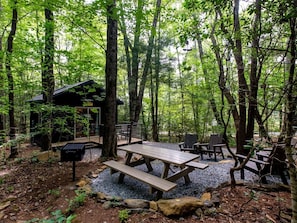 Un terrain de camping boisé, équipé de tables de pique-nique, de chaises, d’un petit chalet et d’un sentier de gravier.