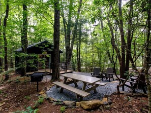 Un campamento arbolado con mesas de picnic, sillas, una pequeña cabaña y un sendero de grava.