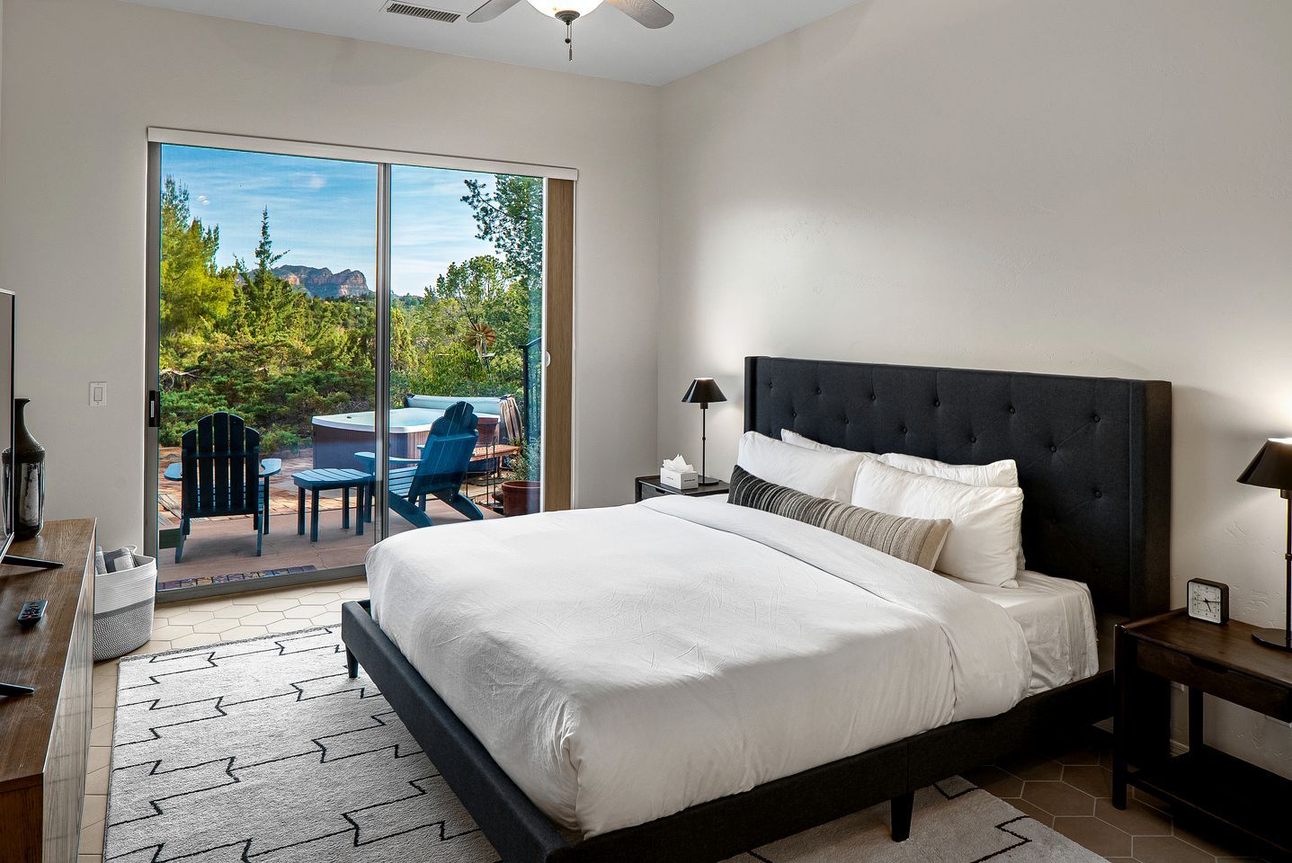 A bedroom with a large bed, a nightstand, a lamp, a desk with a chair, and a sliding glass door leading to an outdoor area with a hot tub and chairs.
