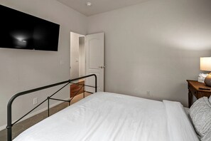 Une chambre à coucher avec un lit, un téléviseur fixé au mur, une table de chevet avec une lampe et une porte donnant sur une autre pièce.