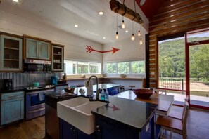 A kitchen with a central island, a sink, and a stove. There are cabinets, a window, and a view of greenery outside. - Stunning Elevated Cabin on the Caddo River for a Peaceful Getaway with Gorgeous Views in Glenwood, Arkansas (Glenwood)