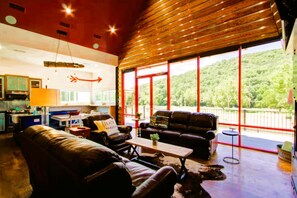 A spacious living area with a large window overlooking a forested landscape, a kitchen area with modern appliances, and a cozy seating arrangement. - Stunning Elevated Cabin on the Caddo River for a Peaceful Getaway with Gorgeous Views in Glenwood, Arkansas (Glenwood)