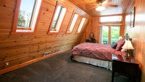 Una habitació amb sostre inclinat, parets de fusta, un llit amb edredó vermell, una cambrera amb una làmpada i una finestra amb vista a vegetació.