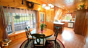 Un interior de madera con una mesa de comedor, sillas, una zona de cocina y un gran ventanal con persianas.