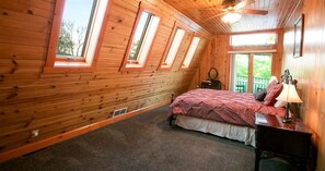 A room with a sloped ceiling, wooden walls, a bed with a red bedspread, a nightstand with a lamp, and a window with a view of greenery. - Romantic Forest Cabin with Amazing Private Deck and Spa in Ohio (Sugarcreek)