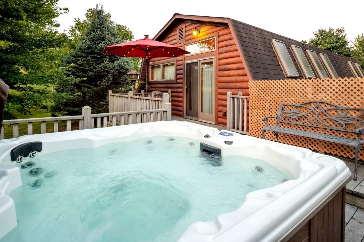 A hot tub in front of a wooden cabin with a deck and a red umbrella.
