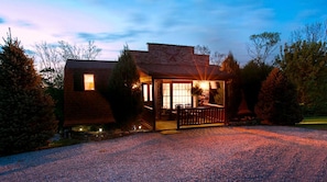 A rustic cabin with a porch, a stone chimney, and a gravel driveway.
