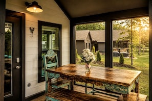 A wooden dining table with chairs, a vase of flowers, and a view of a house and garden through large windows. - Delightful Cabin with Fire Pit and Hot-tub in Tracy City, Tennessee (Tracy City)