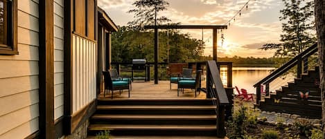 Une terrasse dotée de mobilier d’extérieur, d’un barbecue et offrant une vue sur un lac au coucher du soleil.