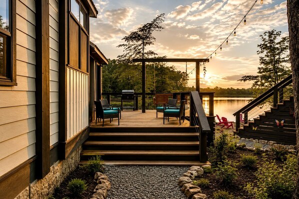 Una terraza con mobiliario exterior, una parrilla y vistas a un lago al atardecer.