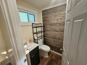 A compact bathroom with a white toilet, a black metal shelf, a white sink, and a wooden wall. - Gorgeous Cottage with Stunning Sights in Wellston, Michigan (Wellston)