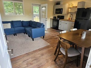 A living room with a blue sectional sofa, a wooden dining table with chairs, a kitchen with white cabinets, a black refrigerator, and a large window.