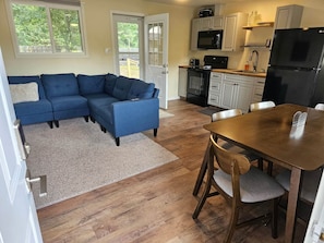 A living room with a blue sectional sofa, a wooden dining table with chairs, a kitchen with white cabinets, a black refrigerator, and a large window. - Gorgeous Cottage with Stunning Sights in Wellston, Michigan (Wellston)