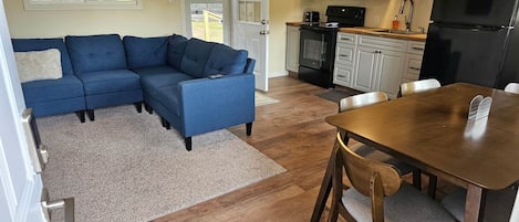 Una sala de estar con un sofá seccional azul, una mesa de comedor de madera con sillas, una cocina con armarios blancos, un refrigerador negro y un ventanal grande.