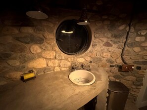 Une salle de bain aux murs en pierre, dotée d’un miroir rond, d’un lavabo et de toilettes.