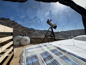 Ein Teleskop auf einem Stativ in einem Raum mit Holzboden, einem Bett und Blick auf eine Berglandschaft durch ein sechseckiges Fenster.